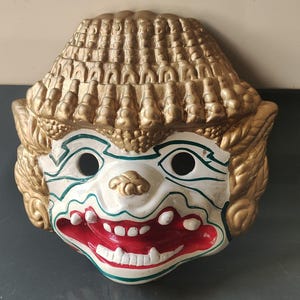 May include: A decorative mask with a white base, featuring a red mouth and teeth, and green and gold accents. The mask has a gold headdress and side details, with two eye holes. The mask has a smiling expression.