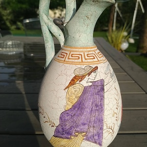 May include: A ceramic pitcher with a green glaze and a handle shaped like a snake. The pitcher is decorated with a painted scene of a woman in a purple robe holding a flower.