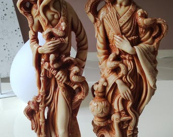 May include: Two decorative figurines of a man and woman in traditional Asian clothing. The figurines are made of a cream-colored material with brown accents and intricate details. The man is holding a staff and the woman is holding a bird.