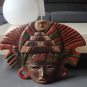 Op de afbeelding: Een gedetailleerd, handgeschilderd Azteeks masker in bruin, rood en groen. Het masker heeft een piramide-ontwerp, een maan en ingewikkelde gravures, wat een cultureel artefact laat zien.
