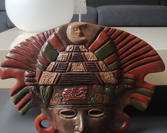May include: A detailed, hand-painted Aztec mask in shades of brown, red, and green. The mask features a pyramid design, a crescent moon, and intricate carvings, showcasing a cultural artifact.