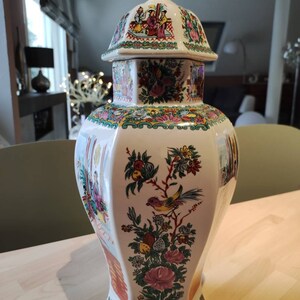 Puede incluir: Un jarrón de cerámica blanca con tapa, que presenta un diseño floral y de pájaros. El jarrón tiene una base hexagonal y un cuerpo redondeado. La tapa está decorada con una escena de personas con ropa tradicional.