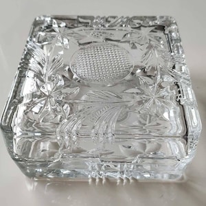 May include: A clear, rectangular glass trinket box with a lid. The box features intricate floral and geometric patterns etched into the glass. The lid has a textured, circular design in the center.