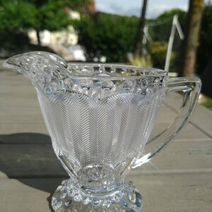 Peut inclure: Un pot à crème en verre transparent avec un motif en chevron texturé et un bord perlé. Le pot à crème a une base ronde avec un motif perlé.