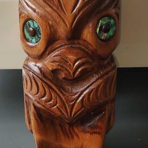 Könnte beinhalten: Eine geschnitzte Tiki-Statue aus Holz mit detailliertem Design. Der Tiki hat eingelegte Augen mit einem grün-blauen, irisierenden Material. Die Statue hat eine warme braune Farbe und eine Zunge, die sich vom Boden erstreckt.