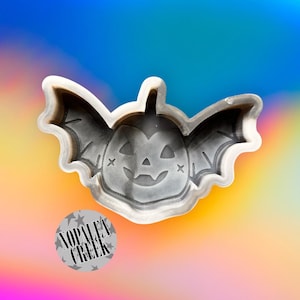 May include: A gray plastic Halloween cookie cutter in the shape of a pumpkin with bat wings. The pumpkin has a smiling face with cross-shaped eyes. The text "NOPALEA CREEK" is on a circular sticker.