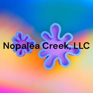 May include: Two purple plastic cookie cutters in the shape of a flower. The larger cutter has a more abstract shape with five points. The smaller cutter has a more traditional flower shape with six points. The background is a blurred rainbow gradient.