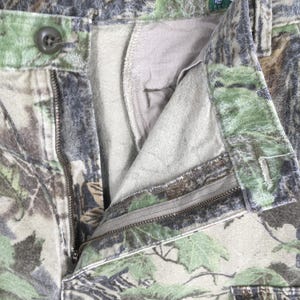May include: Close-up of camouflage pants with an open zipper and button closure. The pants feature a pattern of green, brown, and gray, resembling a forest environment. The interior lining is a light beige color.
