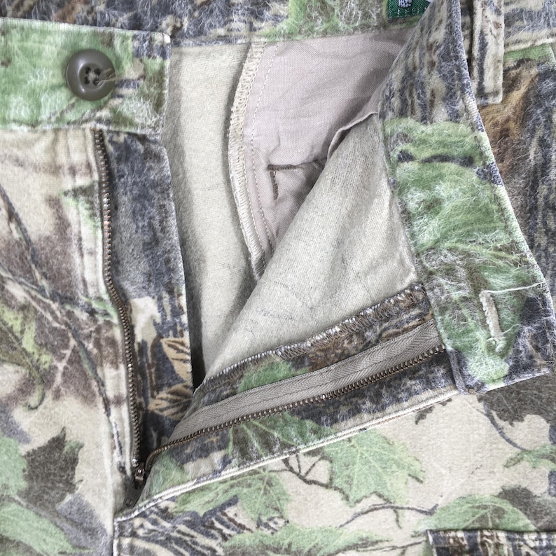 May include: Close-up of camouflage pants with an open zipper and button closure. The pants feature a pattern of green, brown, and gray, resembling a forest environment. The interior lining is a light beige color.