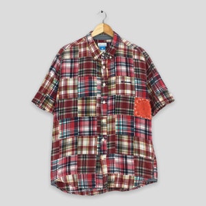 May include: A red, green, and blue plaid short-sleeved button-down shirt with a patchwork design. The shirt has a small red and white bandana patch on the right sleeve and the word "Columbia" embroidered on the left chest.