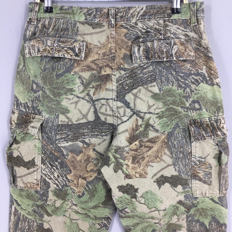 May include: Close-up of camouflage cargo pants. The pants feature a woodland camouflage pattern with shades of green, brown, and tan. The design includes leaf and tree branch details. The pants have multiple pockets.