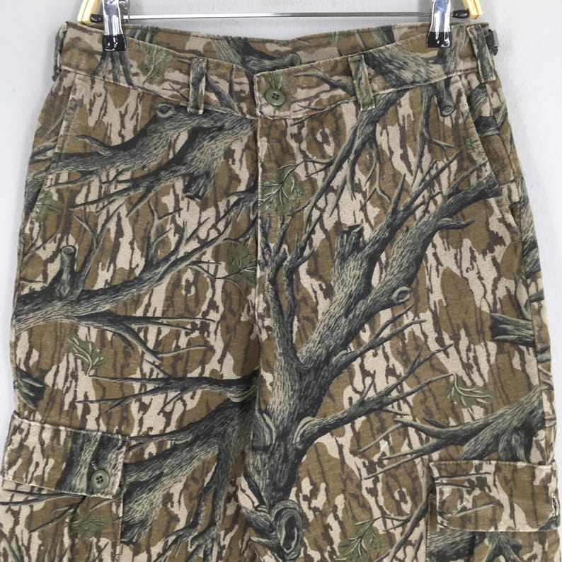 Pu&ograve; includere: Pantaloni cargo mimetici nei toni del marrone, beige e verde, con un motivo a ramo d'albero. I pantaloni hanno una chiusura con bottoni, passanti per cintura e pi&ugrave; tasche. Il tessuto sembra essere una miscela di cotone resistente.