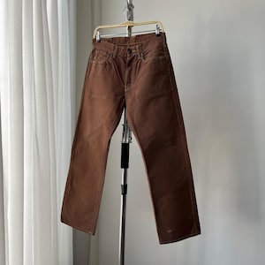 Könnte beinhalten: Braune Jeans aus Denim, die an einer Metallkleiderstange hängen. Die Jeans haben ein klassisches Fünf-Taschen-Design und eine gerade Beinpassform. Die Farbe ist einfarbig mittelbraun, und der Stoff scheint leicht abgenutzt zu sein.