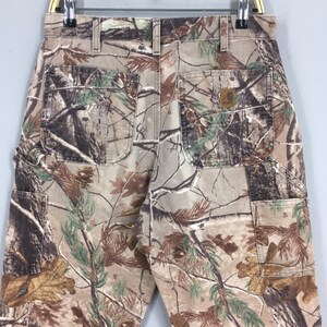 K&ouml;nnte beinhalten: Arbeitshose mit Tarnmuster mit mehreren Taschen und einem Carhartt-Markenaufn&auml;her. Die Hose hat eine Mischung aus braunen, gr&uuml;nen und beigen Farben, die sich in eine Waldumgebung einf&uuml;gen sollen. Die Hose h&auml;ngt an einem Metallb&uuml;gel.