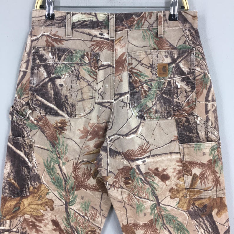 K&ouml;nnte beinhalten: Arbeitshose mit Tarnmuster mit mehreren Taschen und einem Carhartt-Markenaufn&auml;her. Die Hose hat eine Mischung aus braunen, gr&uuml;nen und beigen Farben, die sich in eine Waldumgebung einf&uuml;gen sollen. Die Hose h&auml;ngt an einem Metallb&uuml;gel.