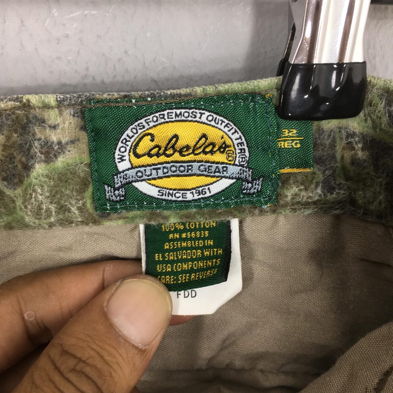 May include: Close-up of a Cabela's outdoor gear tag. The tag is green with gold lettering and features the text "WORLD'S FOREMOST OUTFITTER" and "OUTDOOR GEAR SINCE 1961". The tag also includes size information and fabric details.