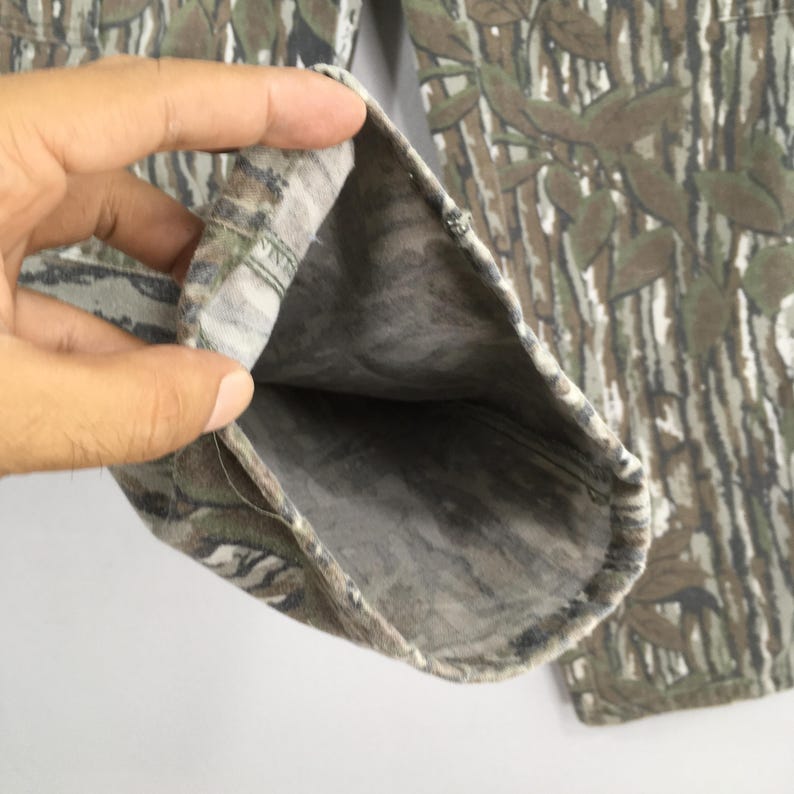 May include: Camouflage-patterned pants with a detailed print of leaves and tree bark in shades of green, brown, and gray. The fabric is folded, revealing the interior lining. The pants are designed for outdoor activities.