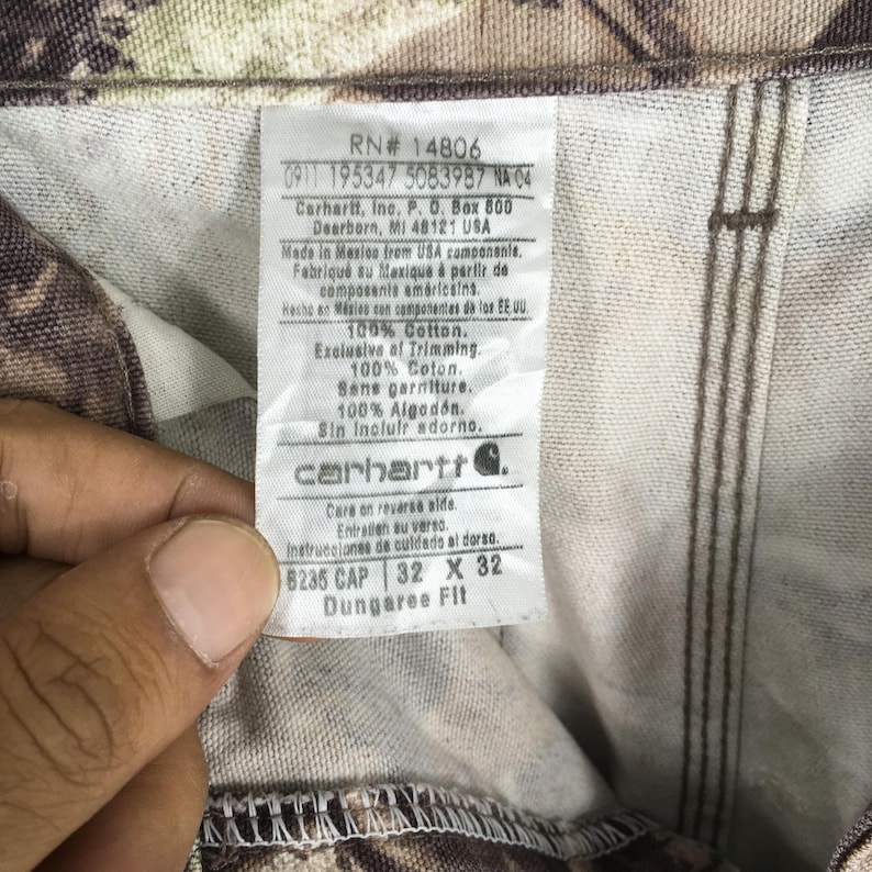 K&ouml;nnte beinhalten: Nahaufnahme einer Carhartt Dungaree Fit Hose mit Camouflagemuster. Das Etikett lautet "8235 CAP 32 X 32" und enth&auml;lt das Carhartt-Logo und Herstellungsdetails. Die Hose besteht aus 100 % Baumwolle.