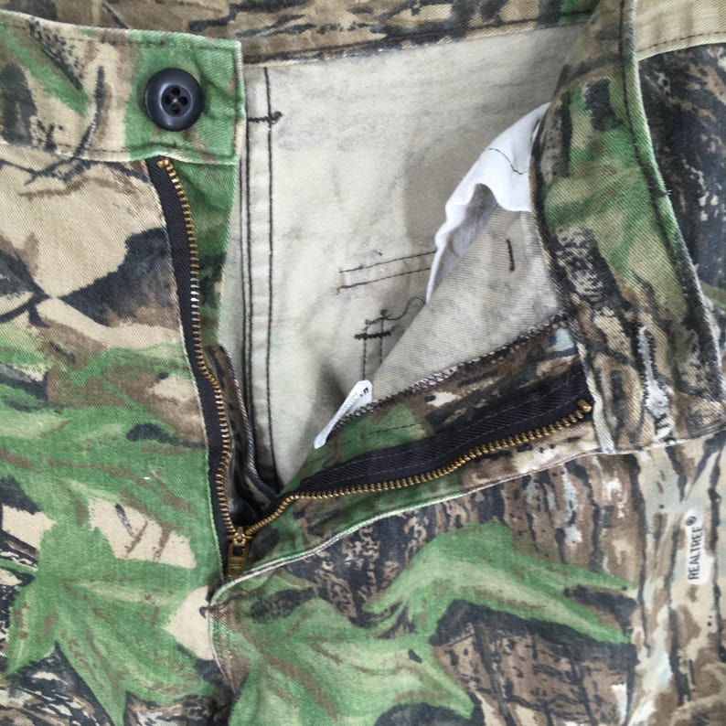 Puede incluir: Primer plano de pantalones de camuflaje con una cremallera negra parcialmente desabrochada. Los pantalones tienen un cierre de bot&oacute;n y una mezcla de colores verdes, marrones y beige. La tela presenta un patr&oacute;n realista de &aacute;rboles y hojas.