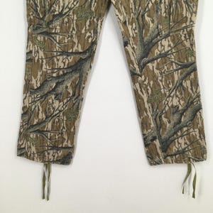 Pu&ograve; includere: Pantaloni mimetici con un motivo dettagliato di rami di alberi nei toni del marrone, verde e beige. I pantaloni hanno un cordoncino sul fondo di ogni gamba. Il tessuto sembra essere un materiale resistente.
