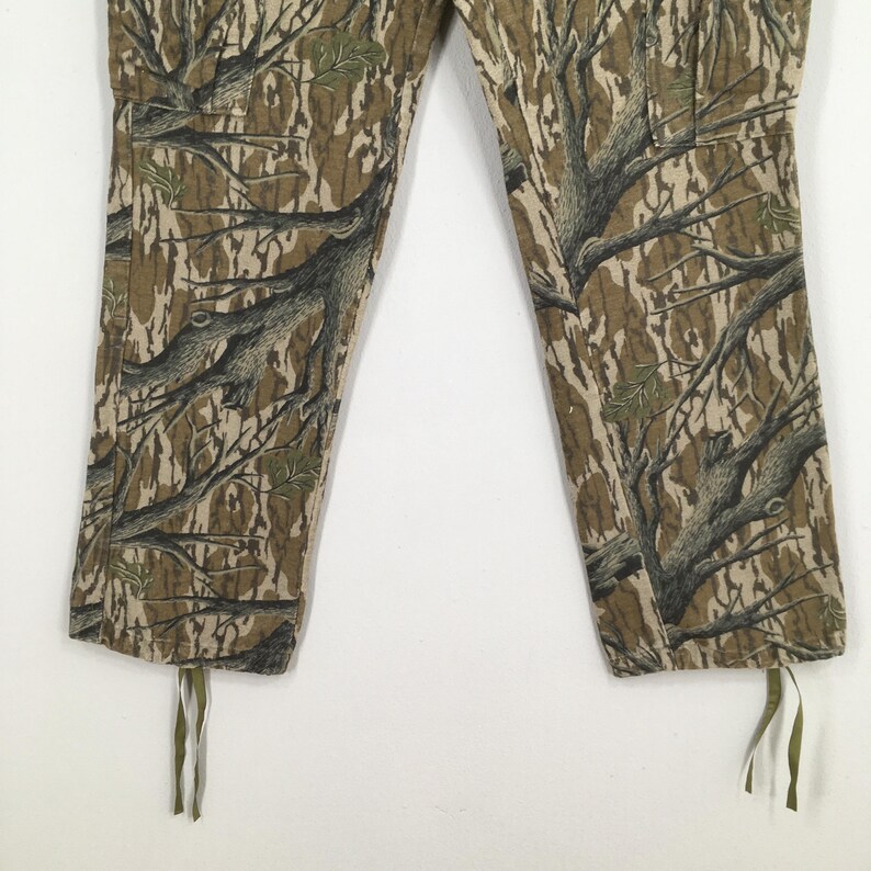 Pu&ograve; includere: Pantaloni mimetici con un motivo dettagliato di rami di alberi nei toni del marrone, verde e beige. I pantaloni hanno un cordoncino sul fondo di ogni gamba. Il tessuto sembra essere un materiale resistente.