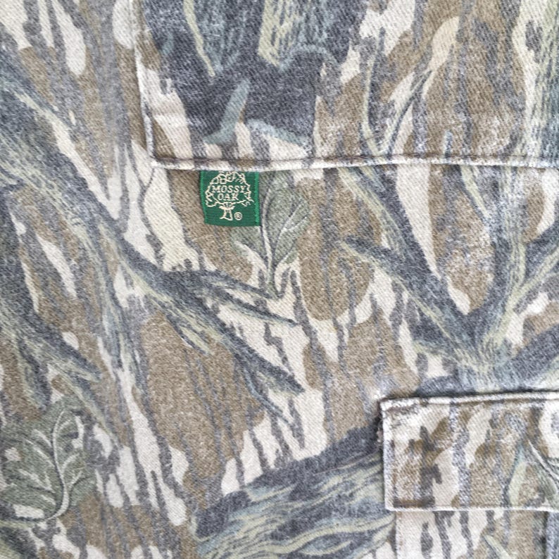 May include: Close-up of a camouflage pattern fabric with two pockets. The fabric features a blend of green, brown, and gray hues, creating a naturalistic design. A green tag with the "Mossy Oak" logo is visible.