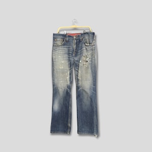 May include: Faded blue denim jeans with distressed details, including rips and paint splatters. The jeans are hanging on a yellow hanger, showcasing a classic five-pocket design and a button-fly closure. The jeans have a straight leg cut.