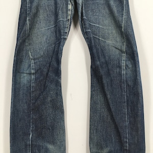 Puede incluir: Primer plano de unos vaqueros de mezclilla azules con un lavado descolorido. Los vaqueros tienen un dise&ntilde;o cl&aacute;sico de cinco bolsillos y una pernera recta. La mezclilla tiene un aspecto desgastado con decoloraci&oacute;n y bigotes sutiles.