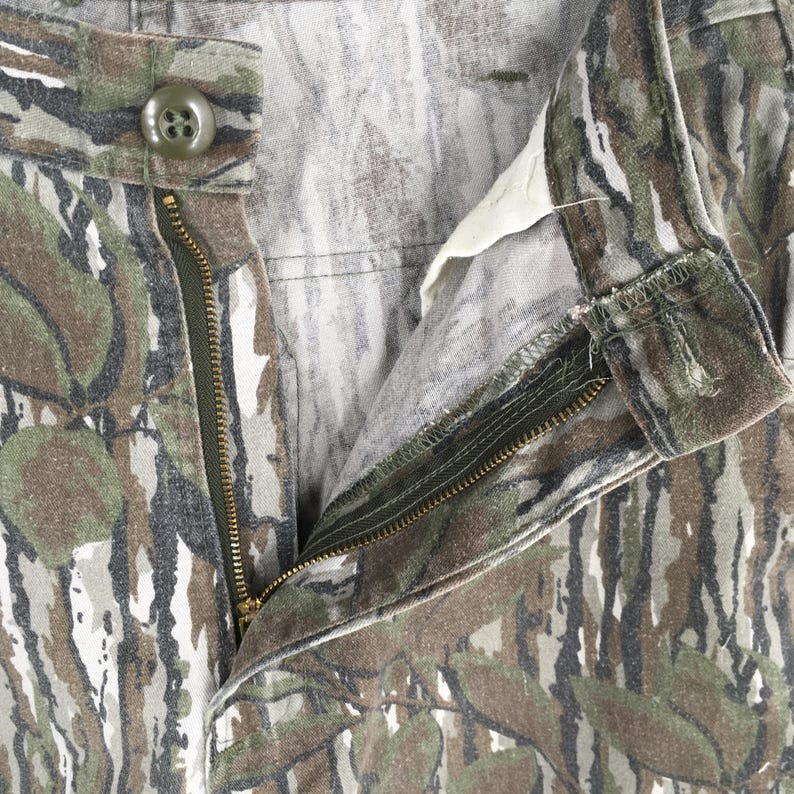 May include: Close-up of camouflage pants with an open zipper and button closure. The pants feature a pattern of green, brown, and gray, resembling a forest or woodland design. The zipper is brass-colored, and the button is olive green.
