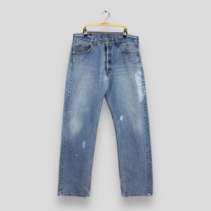 May include: Light blue denim jeans with a button fly, distressed details, and paint splatters. The jeans are hanging on a gold-colored hanger. The jeans have a classic five-pocket design and a straight leg fit.