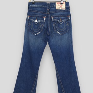 May include: Dark blue denim jeans with a flared leg design. The jeans feature classic five-pocket styling, with decorative stitching on the back pockets and a leather brand patch. The jeans are hanging on a gold-colored hanger.