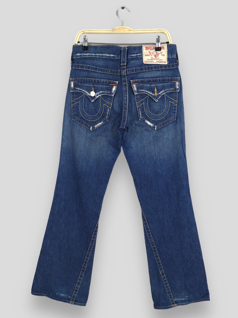 May include: Dark blue denim jeans with a flared leg design. The jeans feature classic five-pocket styling, with decorative stitching on the back pockets and a leather brand patch. The jeans are hanging on a gold-colored hanger.
