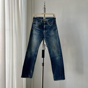 May include: Dark wash denim jeans with a button fly and classic five-pocket design. The jeans are hanging on a metal hanger against a white backdrop. The denim has a faded, worn appearance.