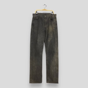 Puede incluir: Un par de vaqueros de mezclilla negros descoloridos cuelgan de una percha de madera. Los vaqueros tienen un corte de pierna recta y un diseño clásico de cinco bolsillos. La tela muestra signos de desgaste y un aspecto sutil y envejecido. Miden unos 100 cm de largo.