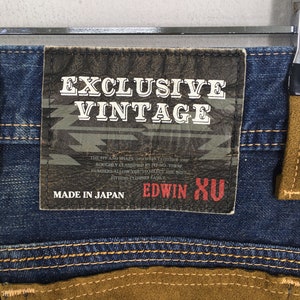 May include: Close-up of blue denim jeans with brown suede-like pocket details. A leather patch reads "EXCLUSIVE VINTAGE" and "MADE IN JAPAN". The jeans have a button with a red center.