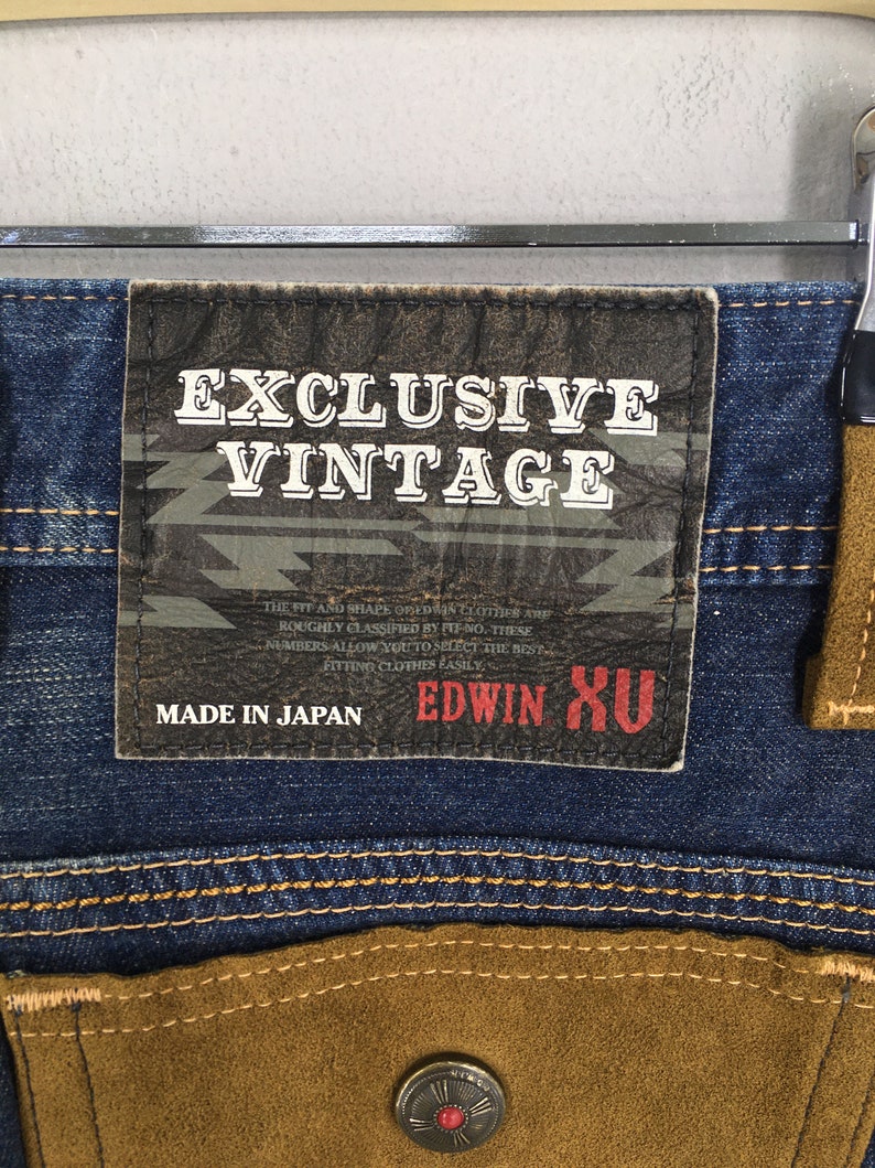 May include: Close-up of blue denim jeans with brown suede-like pocket details. A leather patch reads "EXCLUSIVE VINTAGE" and "MADE IN JAPAN". The jeans have a button with a red center.
