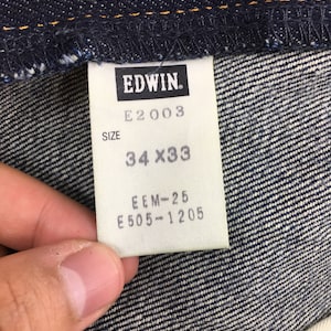 Puede incluir: Primer plano de una tela vaquera azul oscuro con una etiqueta blanca. La etiqueta muestra "EDWIN", "E2003" y la talla "34 x 33". Tambi&eacute;n son visibles los c&oacute;digos "EEM-25" y "E505-1205". El tejido vaquero tiene un aspecto texturizado.