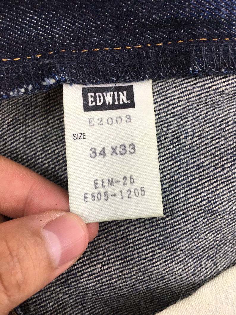 Puede incluir: Primer plano de una tela vaquera azul oscuro con una etiqueta blanca. La etiqueta muestra "EDWIN", "E2003" y la talla "34 x 33". Tambi&eacute;n son visibles los c&oacute;digos "EEM-25" y "E505-1205". El tejido vaquero tiene un aspecto texturizado.
