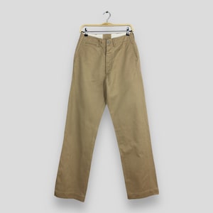 May include: A pair of khaki-colored pants with a button fly and belt loops. The pants are hanging on a gold hanger.