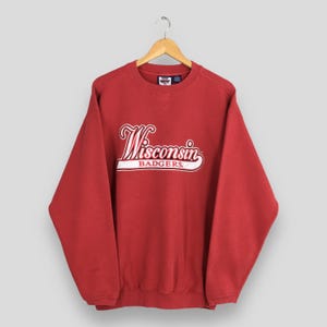 May include: A red sweatshirt with the word "Wisconsin" in a cursive font and the word "Badgers" in a block font. The sweatshirt is hanging on a wooden hanger.