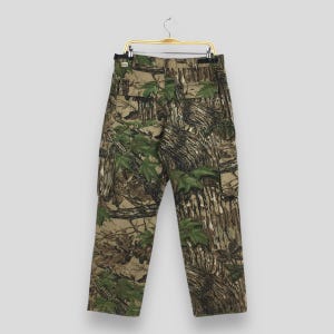 Puede incluir: Pantalones cargo de camuflaje con un corte cl&aacute;sico, con tonos verdes, marrones y beige. Los pantalones tienen m&uacute;ltiples bolsillos y est&aacute;n colgados de una percha met&aacute;lica. La tela muestra un patr&oacute;n forestal realista.