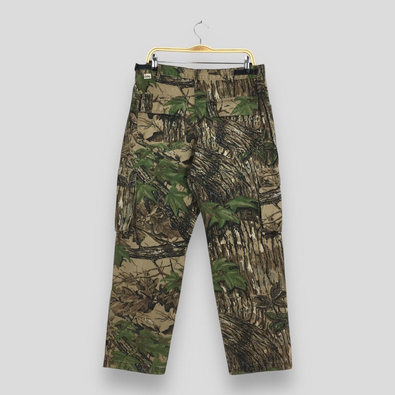 Puede incluir: Pantalones cargo de camuflaje con un corte cl&aacute;sico, con tonos verdes, marrones y beige. Los pantalones tienen m&uacute;ltiples bolsillos y est&aacute;n colgados de una percha met&aacute;lica. La tela muestra un patr&oacute;n forestal realista.
