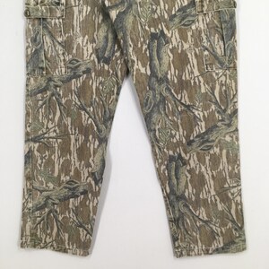 May include: Camouflage cargo pants in shades of brown, green, and tan. The pants feature a detailed tree and branch pattern, with multiple pockets and a button closure. The fabric appears to be a durable material.