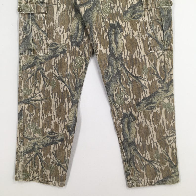 May include: Camouflage cargo pants in shades of brown, green, and tan. The pants feature a detailed tree and branch pattern, with multiple pockets and a button closure. The fabric appears to be a durable material.