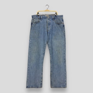 May include: A pair of light blue denim jeans hanging on a wooden hanger. The jeans have a button fly and five pockets.