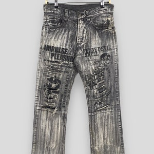 May include: Black distressed denim jeans with a faded white wash. The jeans have multiple rips and tears and feature printed text that says "Impulse Pleasure" and "Funky Devil Skull".