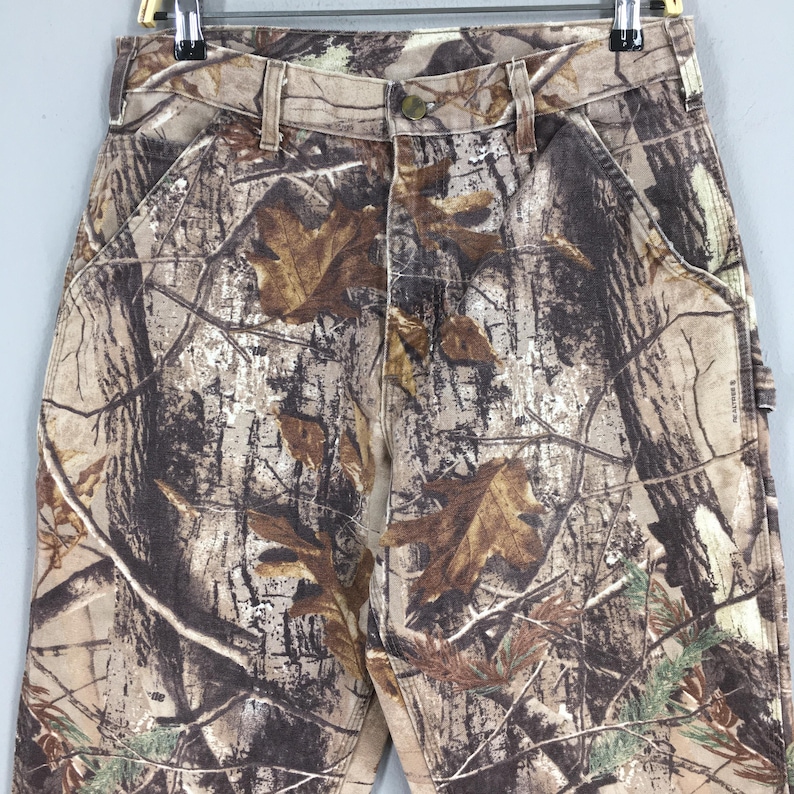 K&ouml;nnte beinhalten: Nahaufnahme einer Camouflage-Hose in Braun-, Beige- und Gr&uuml;nt&ouml;nen. Die Hose hat einen Knopfverschluss, G&uuml;rtelschlaufen und Vordertaschen. Das Tarnmuster umfasst Blatt- und Astdesigns.