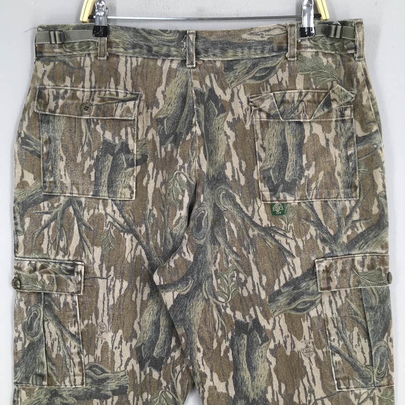 May include: Camouflage cargo pants with multiple pockets. The pants feature a pattern of brown, green, and tan, resembling a forest. The pants are hanging on a hanger.