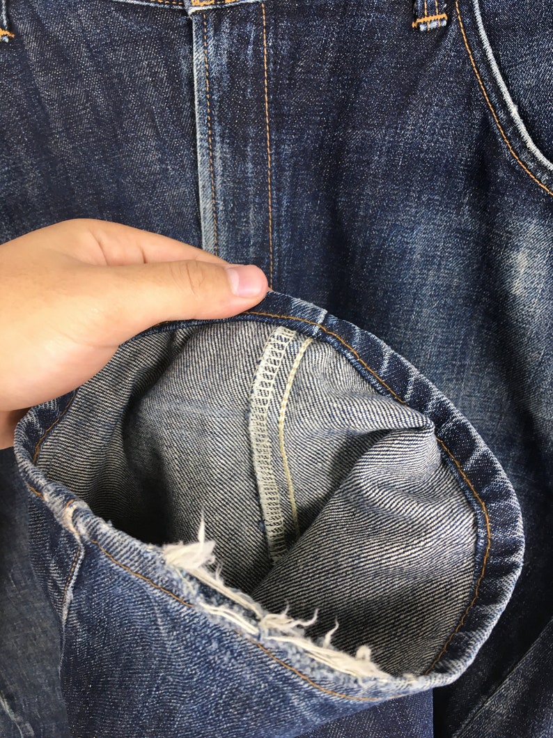 Puede incluir: Primer plano de unos vaqueros de mezclilla azules, que muestran la textura de la tela y los detalles de las costuras. El dobladillo est&aacute; doblado, revelando el interior de la mezclilla. Los vaqueros tienen un aspecto cl&aacute;sico y desgastado.
