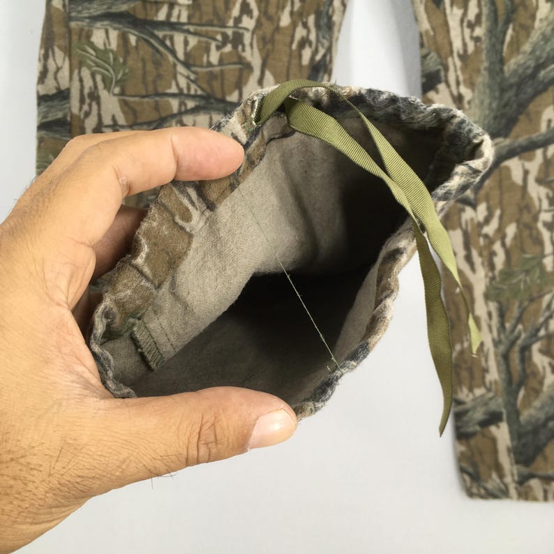 Pu&ograve; includere: Pantaloni mimetici con dettaglio coulisse. I pantaloni presentano un motivo boschivo nei toni del marrone, verde e beige. Il polsino dei pantaloni &egrave; visibile, mostrando la fodera interna e un cordino verde.